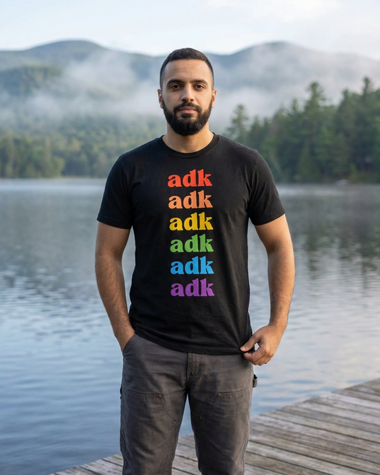 Man wearing a black t-shirt with 'adk' in rainbow colors standing on a dock by a lake.