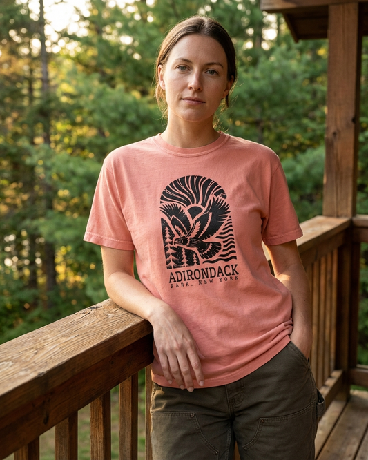 Person wearing a pink t-shirt with Adirondack Park logo on a wooden deck with trees in the background