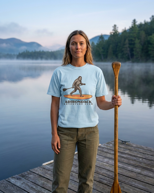 Person standing on a dock by a lake, holding a paddle, wearing a t-shirt with Adirondack Adventure Club design.
