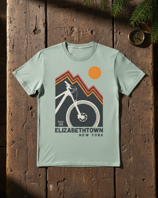 Light green t-shirt with bicycle graphic and 'Elizabethtown, New York' text on a wooden surface.