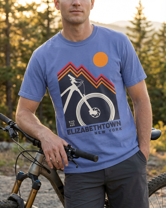 Person wearing a blue t-shirt with a mountain bike graphic and 'Elizabethtown, New York' text, standing next to a bicycle in a forest setting.
