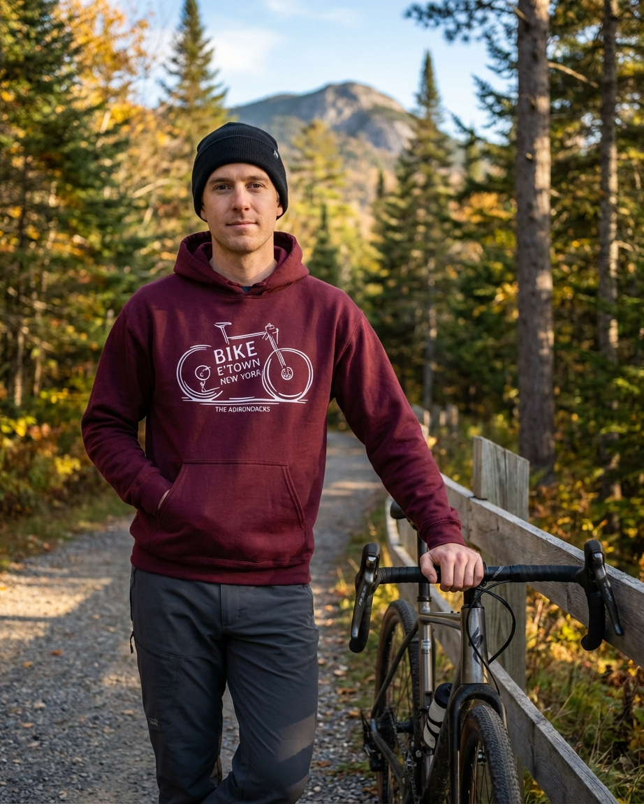Man wearing a maroon hoodie with a bicycle graphic, standing next to a bike on a trail with trees and mountains in the background.