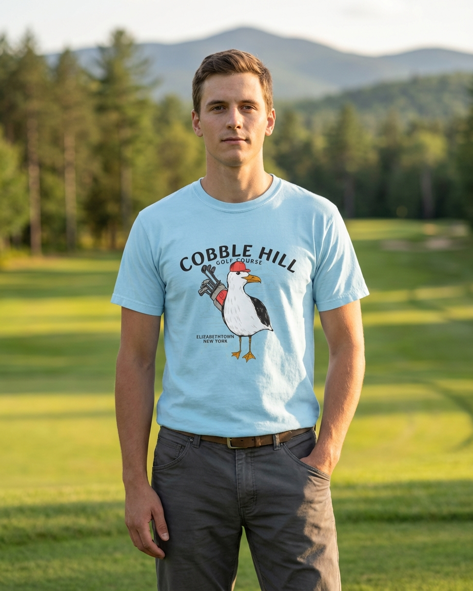 Man wearing a light blue t-shirt with a graphic design of a rooster and text, standing in a grassy field with trees and mountains in the background.