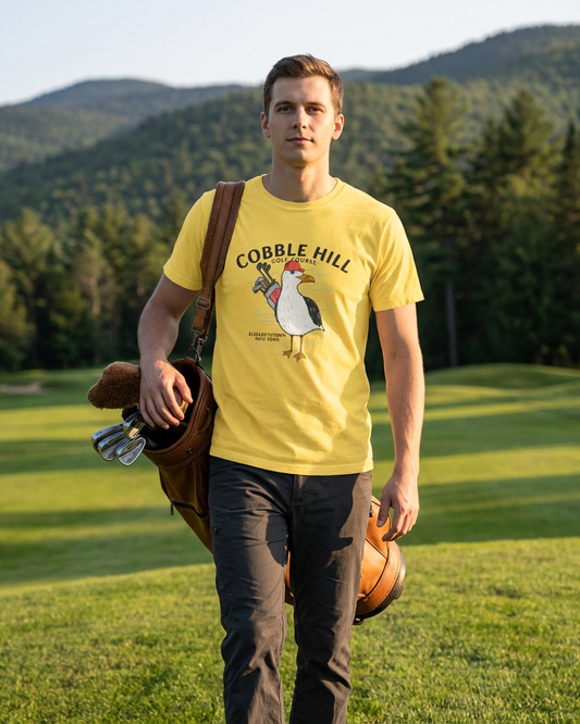 Man wearing a yellow t-shirt with a chicken graphic and text, standing on a golf course with mountains in the background.