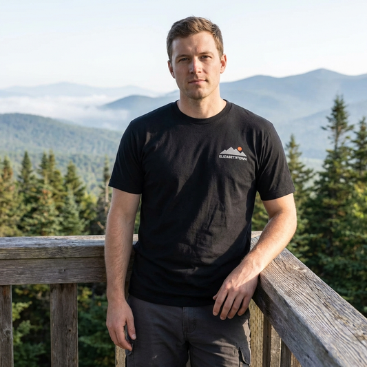 Man wearing a black t-shirt with a logo, standing on a wooden deck with mountains in the background.