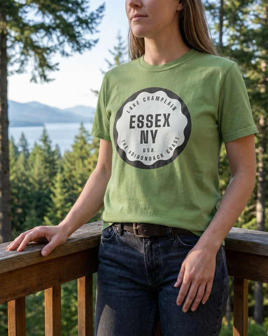 Person wearing a green t-shirt with 'Essex NY' print, standing outdoors with trees and water in the background