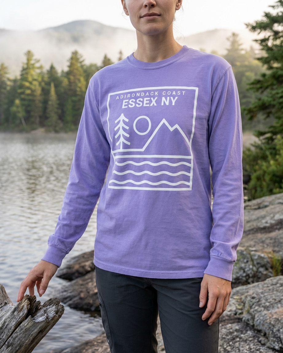 Person wearing a purple long-sleeve shirt with a graphic design standing by a lake with mountains in the background.