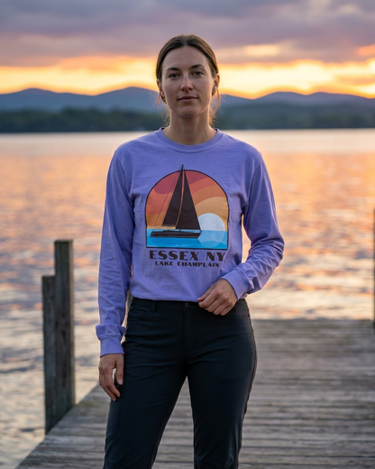 Person wearing a purple sweatshirt with a sailboat design and text, standing on a dock by a lake at sunset.