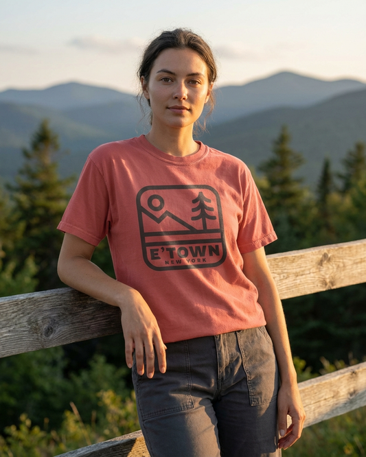 Person wearing a pink t-shirt with a logo and text, standing outdoors with mountains in the background