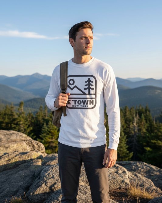 Man standing on a mountain top wearing a white long-sleeve shirt with a logo, holding a backpack.