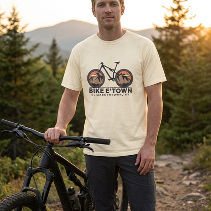 Man wearing a 'Bike E'Town' t-shirt with a mountain bike graphic, standing outdoors with a mountain bike and nature in the background.