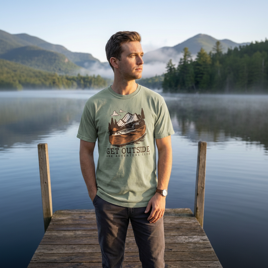 Man standing on a dock by a lake with mountains in the background, wearing a t-shirt with a nature graphic and text.