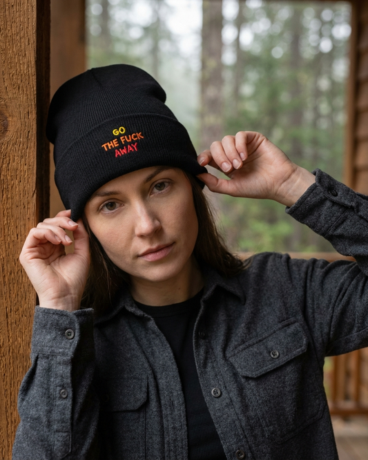 Person wearing a black beanie with "go the fuck away" text, standing in a forested area.