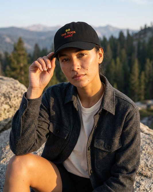 Person wearing a black cap with "go the fuck away" text, sitting outdoors with mountains and trees in the background