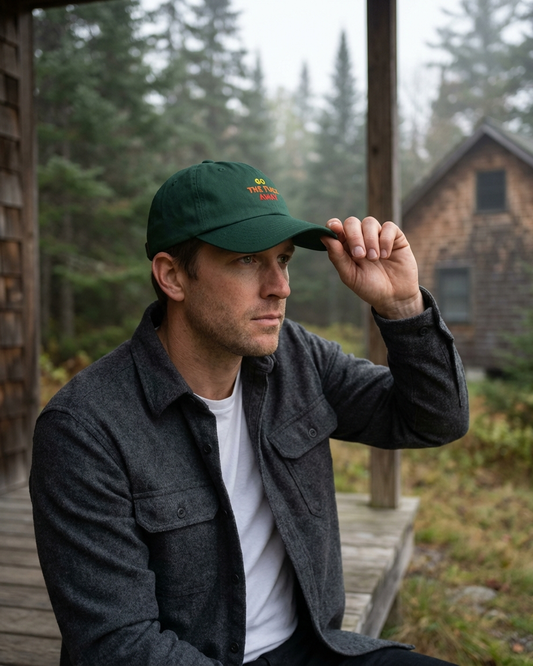 Man wearing a green "Go the fuck away" cap and dark jacket sitting on a wooden porch with a forest background