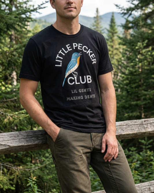Person wearing a black t-shirt with 'Little Pecker Club' design in a forest setting