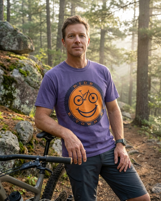 Man wearing a purple t-shirt with a smiley face and bicycle graphic, standing in a forest.
