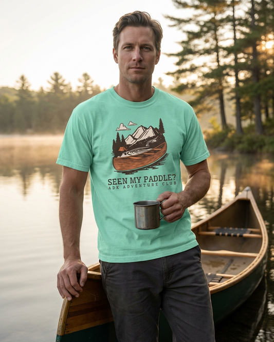 Man wearing a teal t-shirt with a nature graphic and text, standing by a lake with a canoe.