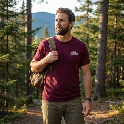 Man in a maroon t-shirt with a logo, holding a backpack, standing in a forest.